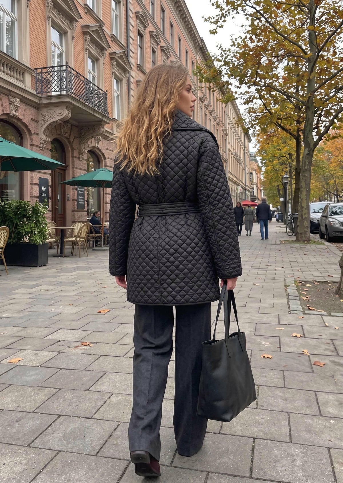 A woman walking on a city sidewalk wearing a black quilted belted coat, dark wide-leg trousers, and carrying a black tote bag.