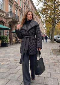 A woman walking on a city sidewalk wearing a black quilted belted coat over a black turtleneck, dark wide-leg pants, and carrying a black tote bag.