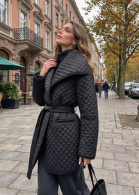 A woman wearing a black quilted belted coat stands on a city sidewalk next to a street with buildings and trees.