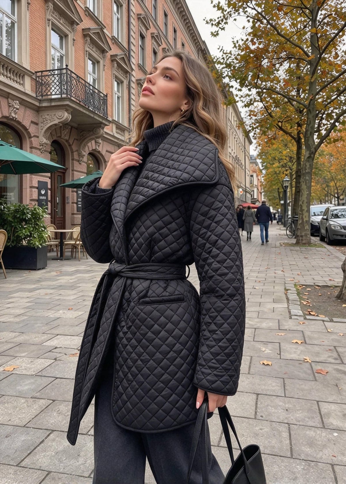 A woman wearing a black quilted belted coat stands on a city sidewalk next to a street with buildings and trees.