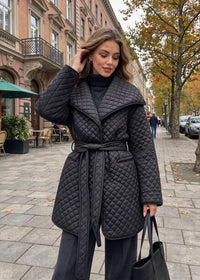 A woman stands on a city sidewalk wearing a black quilted wrap coat with a tie belt, dark wide-leg pants, and holding a black tote bag. The background shows a European-style street with brick buildings, trees, and outdoor cafe seating.