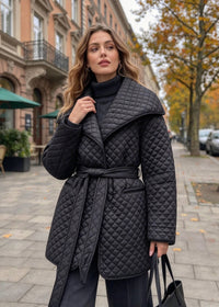 A woman wearing a black quilted belted coat over a black turtleneck sweater, standing on a city sidewalk with buildings and trees in the background.