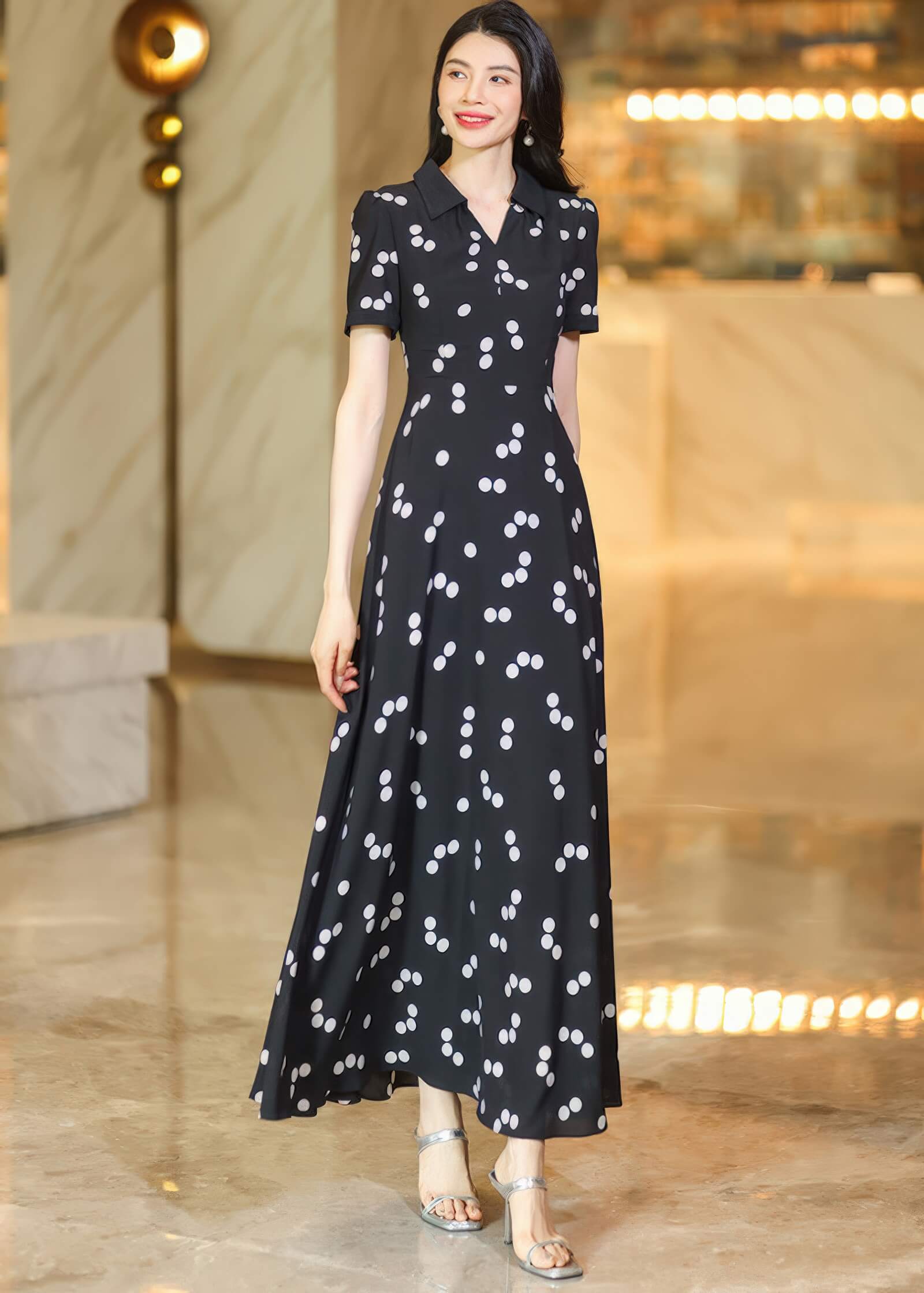 Full-length front view of a woman in a black and white polka dot maxi dress featuring long sleeves and a sophisticated lapel collar.