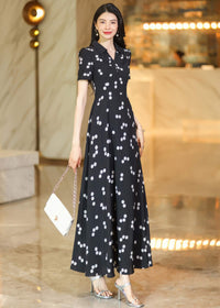 A woman modeling a polka dot maxi dress, accessorized with a small white handbag and silver strappy high heels.