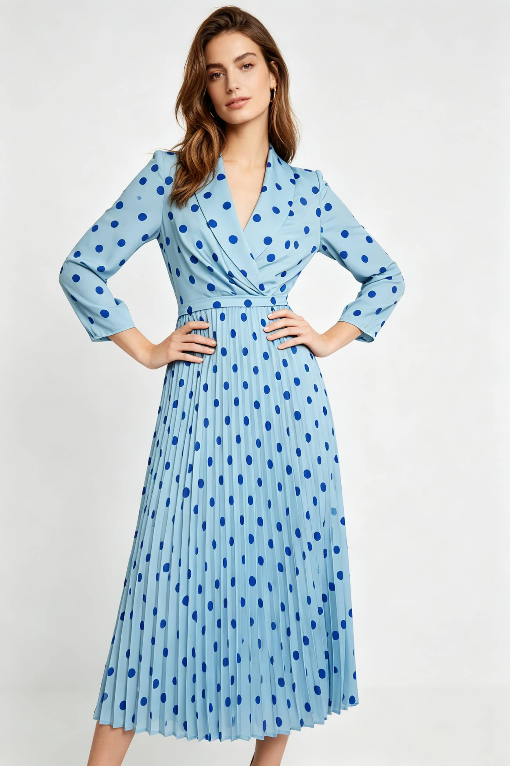 A front-facing studio portrait of a woman standing with both hands on her hips. She is wearing a light blue, wrap-front midi dress with dark blue polka dots, long sleeves, and a structured pleated skirt against a solid white backdrop.