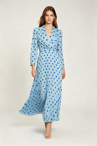 A full-length studio shot of a woman walking forward, causing the pleated skirt of her light blue, dark blue polka-dotted dress to flare slightly. The dress has a wrap-style top, long sleeves, and she is wearing beige high-heeled sandals.