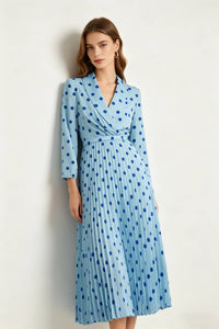 A studio photograph of a woman leaning against a white wall, looking toward the camera. She is wearing a light blue midi dress with dark blue polka dots, a wrap-style bodice with a notched lapel, and long sleeves.