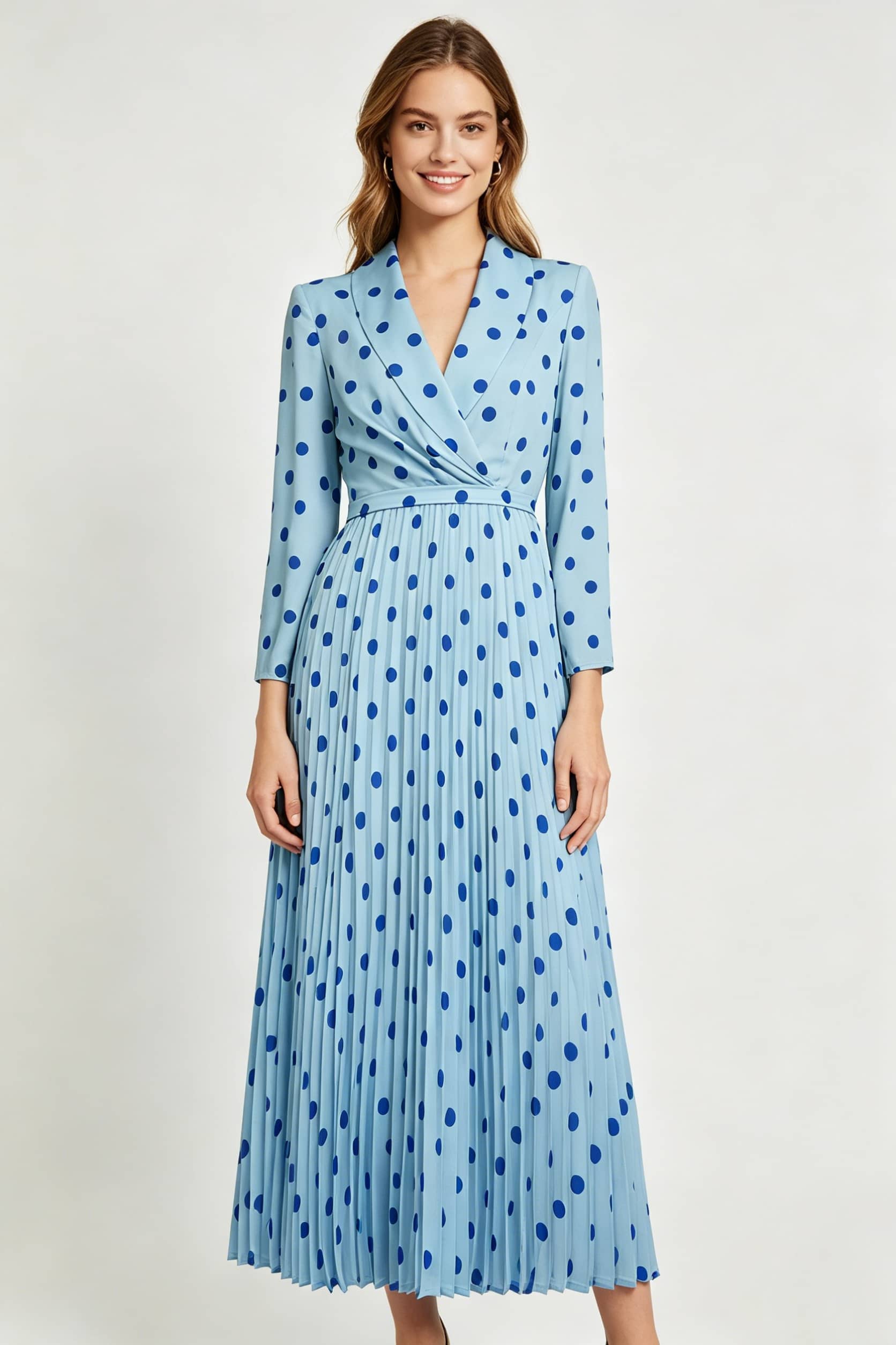 A full-length, front-facing studio shot of a smiling woman standing with her arms at her sides. She is wearing a light blue, long-sleeved polka-dot dress with a wrap front and a long pleated skirt against a plain white background.