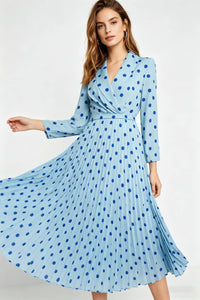A dynamic studio photo of a woman turning to her side, holding out the skirt of her light blue and dark blue polka-dot dress to showcase its full, pleated volume against a white backdrop.