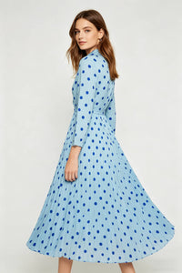 A studio shot capturing a woman from behind as she looks back over her shoulder at the camera. The movement highlights the wide sweep of her pleated, light blue midi dress adorned with dark blue polka dots.