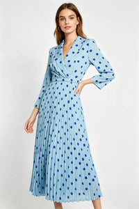 A studio photograph of a woman with brown hair facing slightly left, wearing a light blue, long-sleeved midi dress featuring a dark blue polka-dot pattern, a wrap-style V-neckline with lapels, and a pleated skirt. She has one hand on her hip against a plain white background.