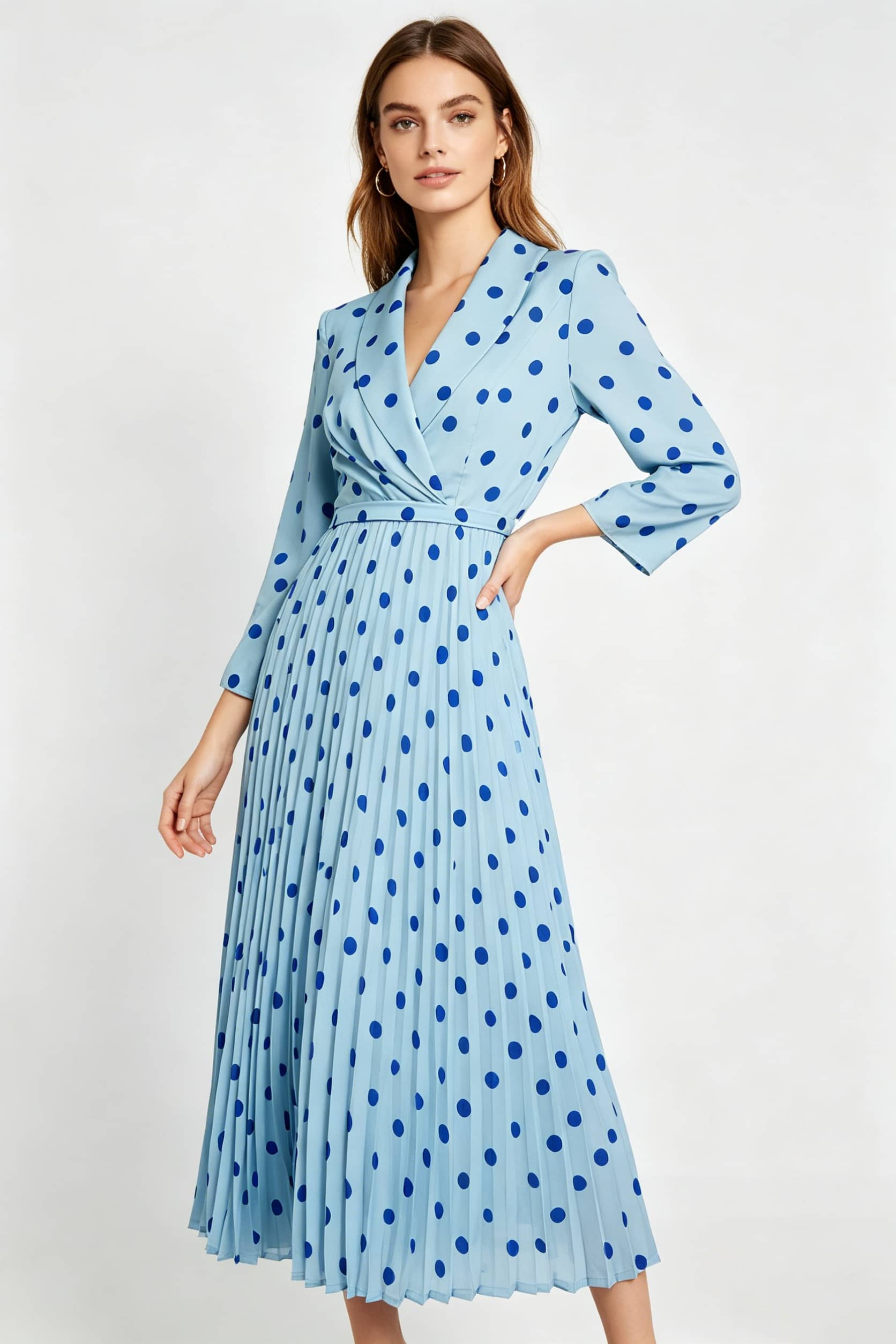A studio photograph of a woman with brown hair facing slightly left, wearing a light blue, long-sleeved midi dress featuring a dark blue polka-dot pattern, a wrap-style V-neckline with lapels, and a pleated skirt. She has one hand on her hip against a plain white background.