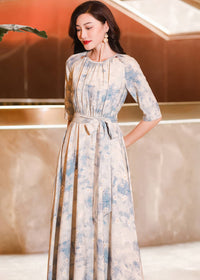 Close-up three-quarter view of a woman looking down, wearing a cream maxi dress with a blue watercolor floral print, gathered neckline, and a matching fabric belt tied at the waist.