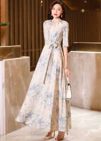 Full-length front view of a woman posing in a cream and blue watercolor floral maxi dress, holding a small white handbag while wearing metallic high heels.