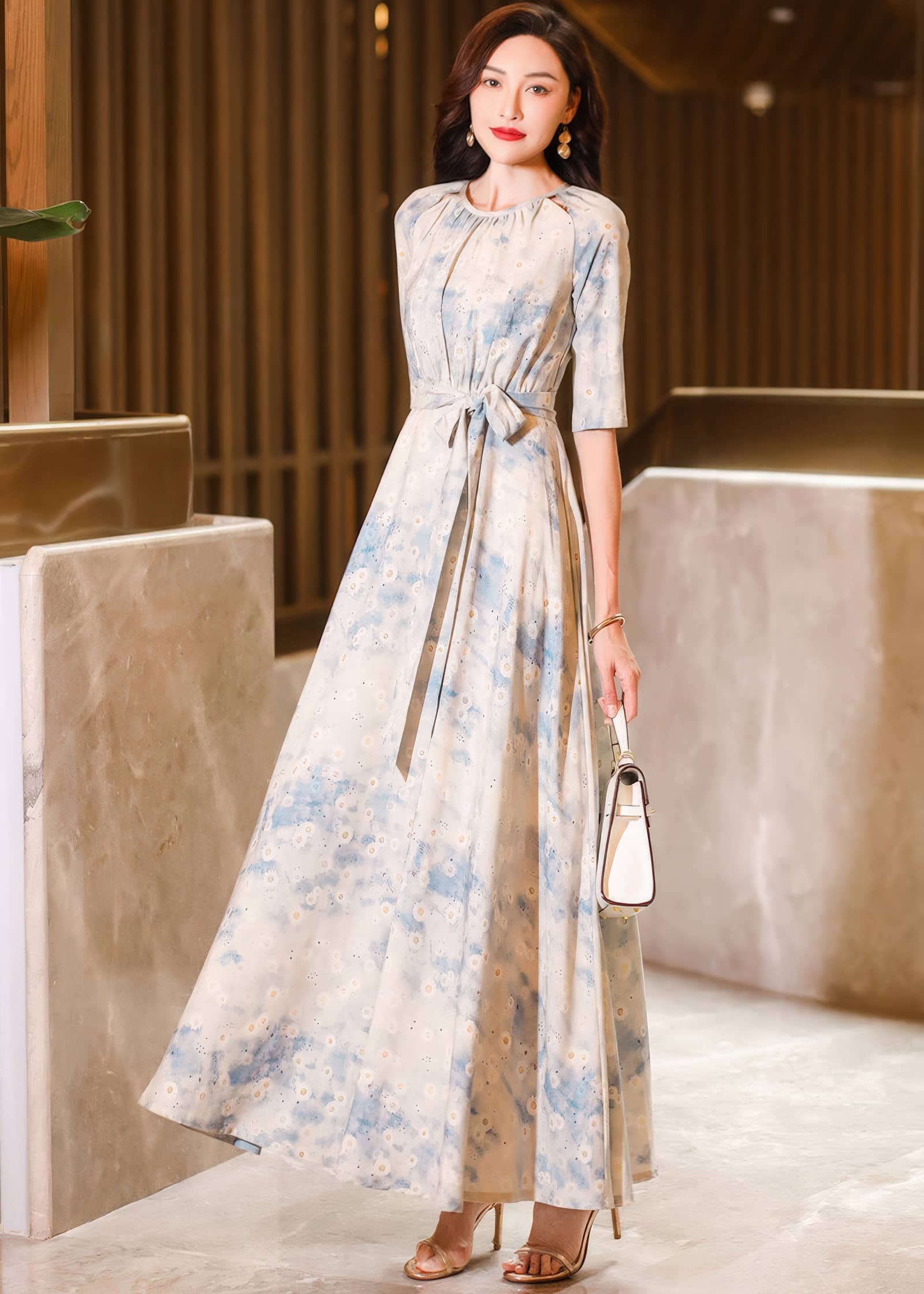 Full-length front view of a woman posing in a cream and blue watercolor floral maxi dress, holding a small white handbag while wearing metallic high heels.