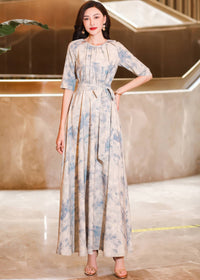 A woman standing with one hand on her hip, modeling a cream-colored, elbow-sleeve maxi dress featuring a soft blue watercolor floral pattern and a tied waist belt.