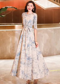 Three-quarter length shot of a woman looking toward the camera in a flowy cream maxi dress with light blue watercolor floral details and a tied waist sash.