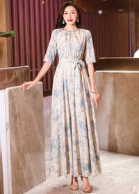 Full-length front view of a woman leaning against a marbled counter structure, wearing a blue watercolor floral maxi dress and gold strappy sandals.