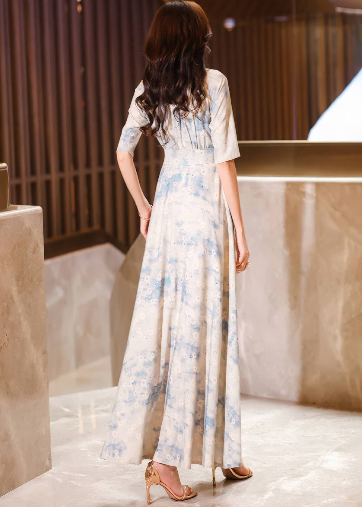 Rear view of a woman with long, wavy brown hair looking over her shoulder, showcasing the back design and floor-length drape of a cream and blue watercolor floral maxi dress.
