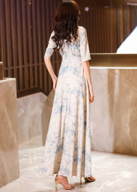 Rear view of a woman with long, wavy brown hair looking over her shoulder, showcasing the back design and floor-length drape of a cream and blue watercolor floral maxi dress.
