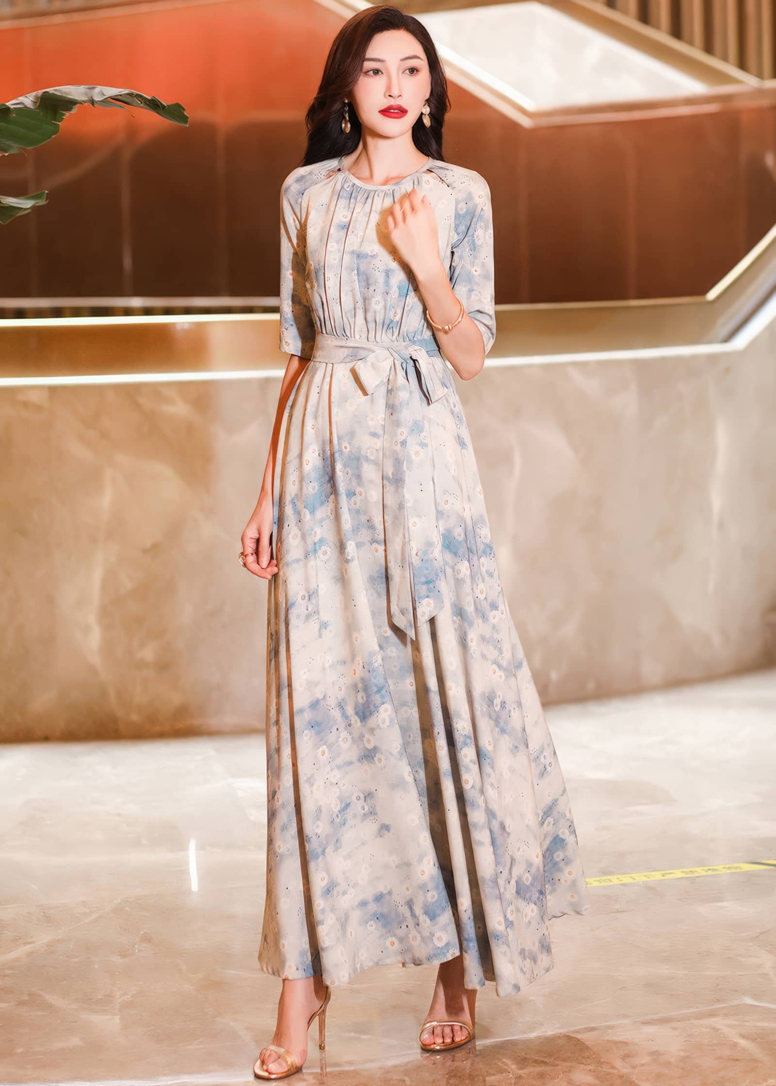 Full-length portrait of a woman posing with one hand near her chest, wearing a cream maxi dress with a soft blue watercolor floral design and a matching tie belt.