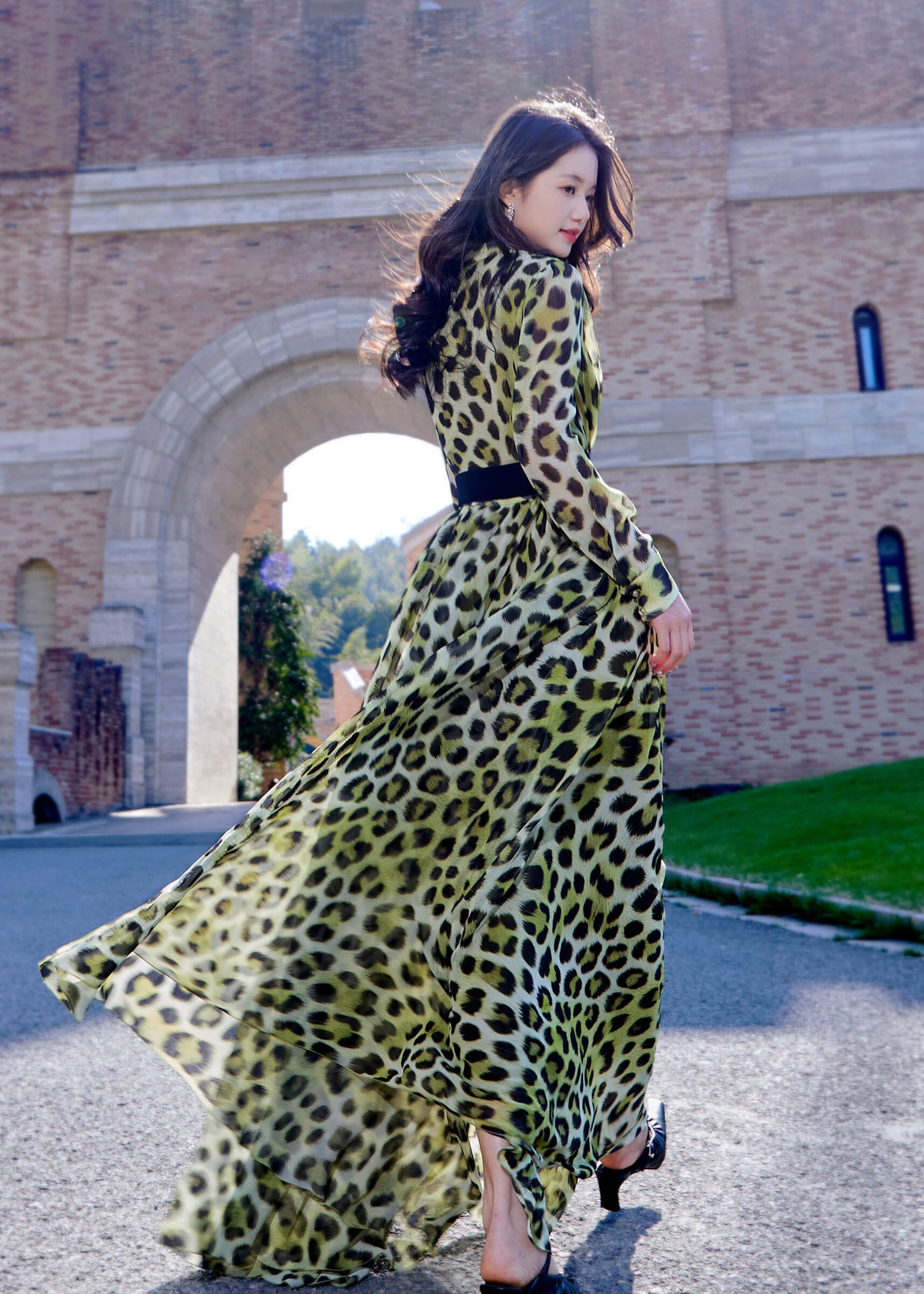 Rear view of a model walking in a floor-length Vivian Seven leopard print maxi dress, showing the dramatic flow of the sheer chiffon fabric under a stone archway.
