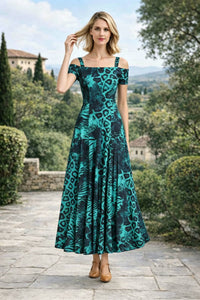 Front view of a woman with one hand in her hair, modeling an emerald green and black leopard print off-the-shoulder A-line maxi dress with thick straps, standing on an outdoor stone patio.