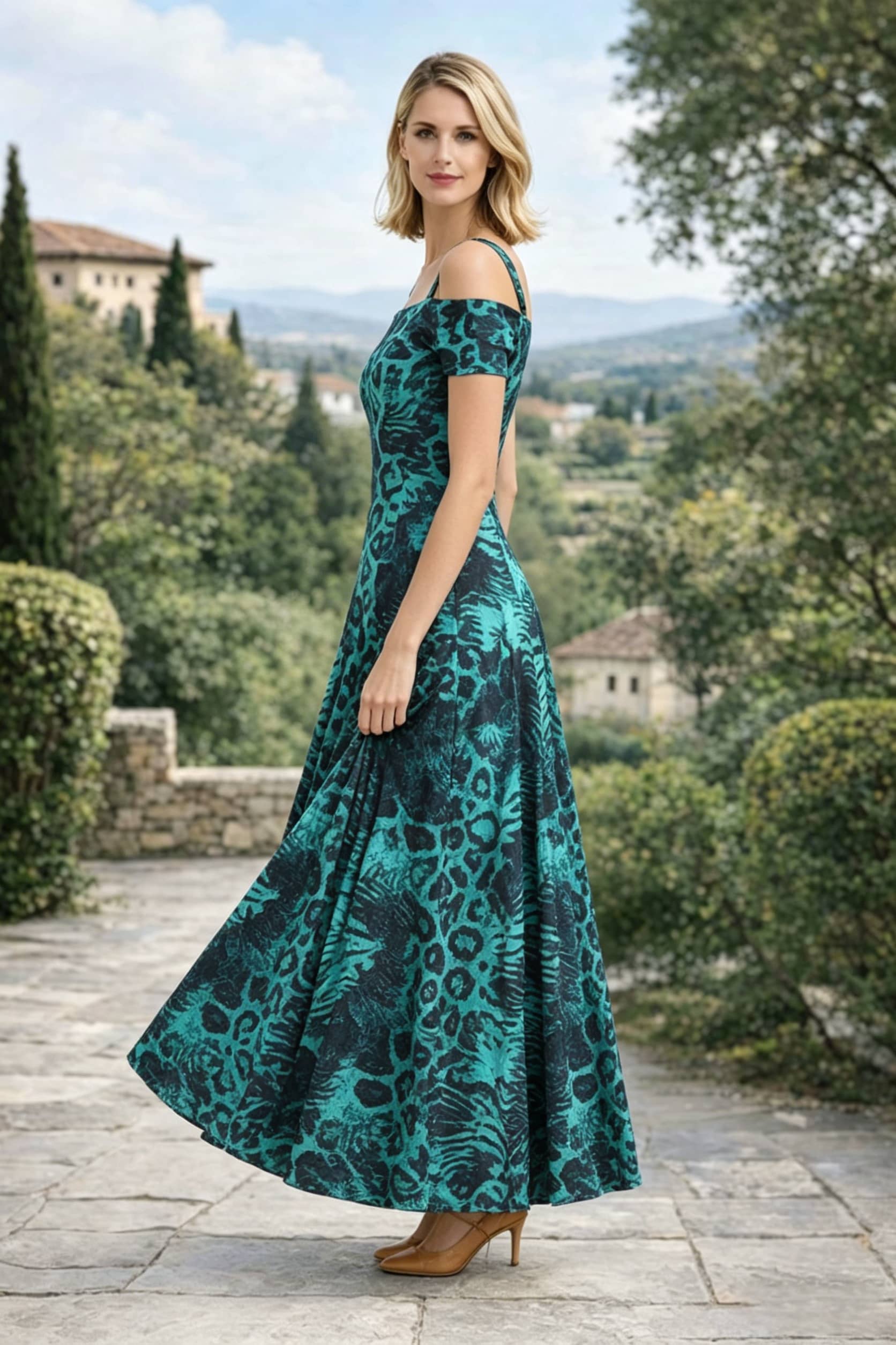 Side profile view of a woman standing on a stone pathway wearing an emerald green and black leopard print off-the-shoulder A-line maxi dress, holding the fabric of the skirt against a scenic European landscape backdrop.