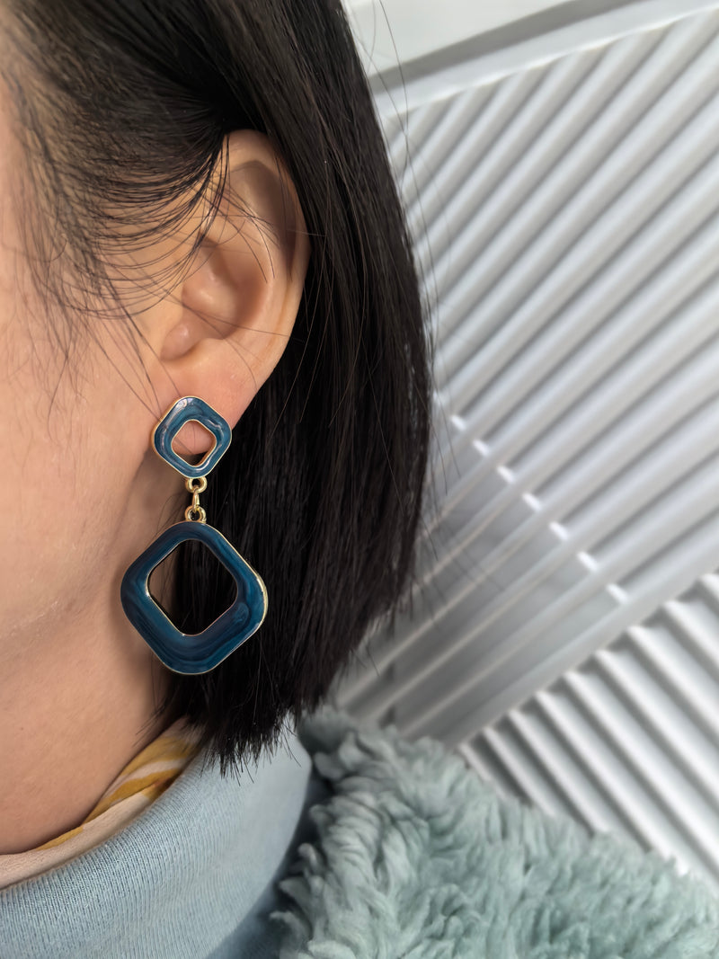 A person wearing the blue enamel version of the earrings, which are made of two connected squares. The earrings dangle from their earlobe, complementing their dark hair and light blue jacket.