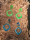 Both the green and blue enamel versions of the earrings are displayed on a bed of shredded brown paper, showcasing their gold-tone finish and two-square design.