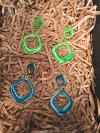 Both the green and blue enamel versions of the earrings are displayed on a bed of shredded brown paper, showcasing their gold-tone finish and two-square design.
