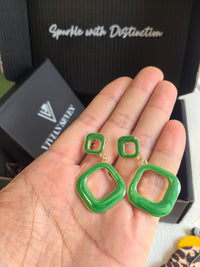 A pair of green enamel earrings with a gold-tone base being held in the palm of a hand. The earrings are made of two connected squares, the smaller one at the top and the larger one dangling below.