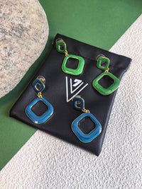 Two pairs of earrings, one in green enamel and one in blue enamel, resting on a black jewelry pouch. The earrings have a modern design with two connected squares and a gold-tone finish.