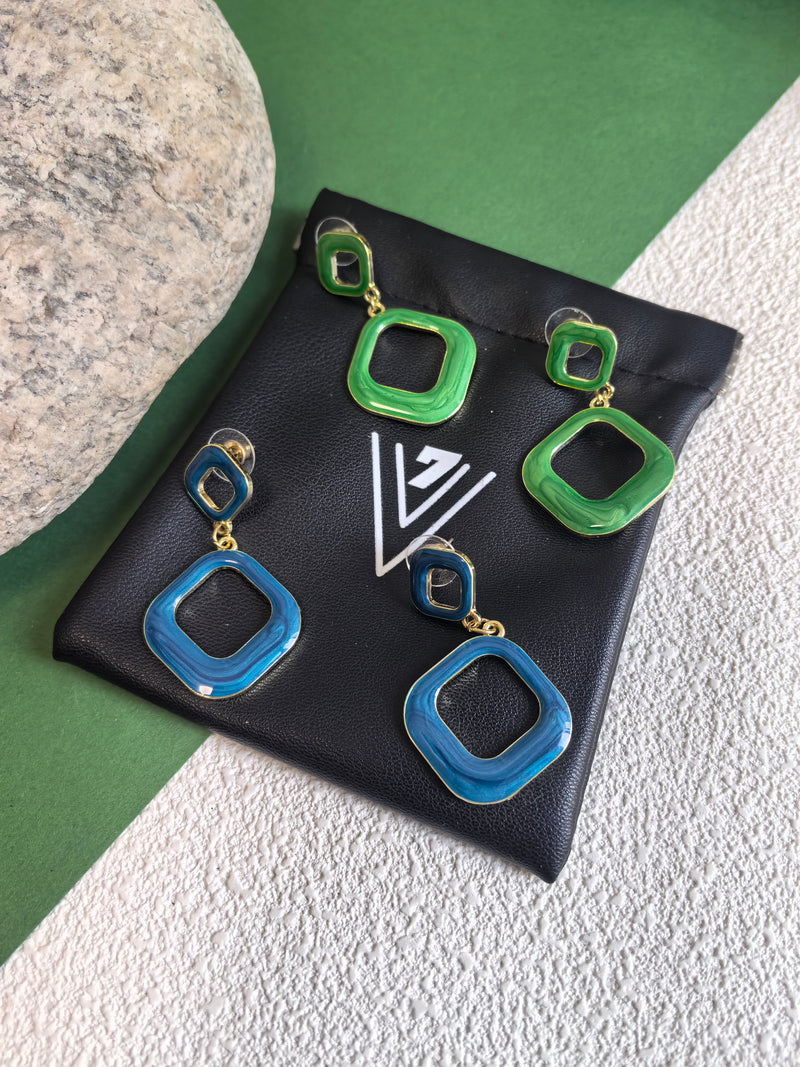 Two pairs of earrings, one in green enamel and one in blue enamel, resting on a black jewelry pouch. The earrings have a modern design with two connected squares and a gold-tone finish.