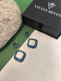 A pair of blue enamel earrings displayed on a beige and green background. The earrings are made of two connected squares and are next to a black box with the brand name "VIVIAN SEVEN."