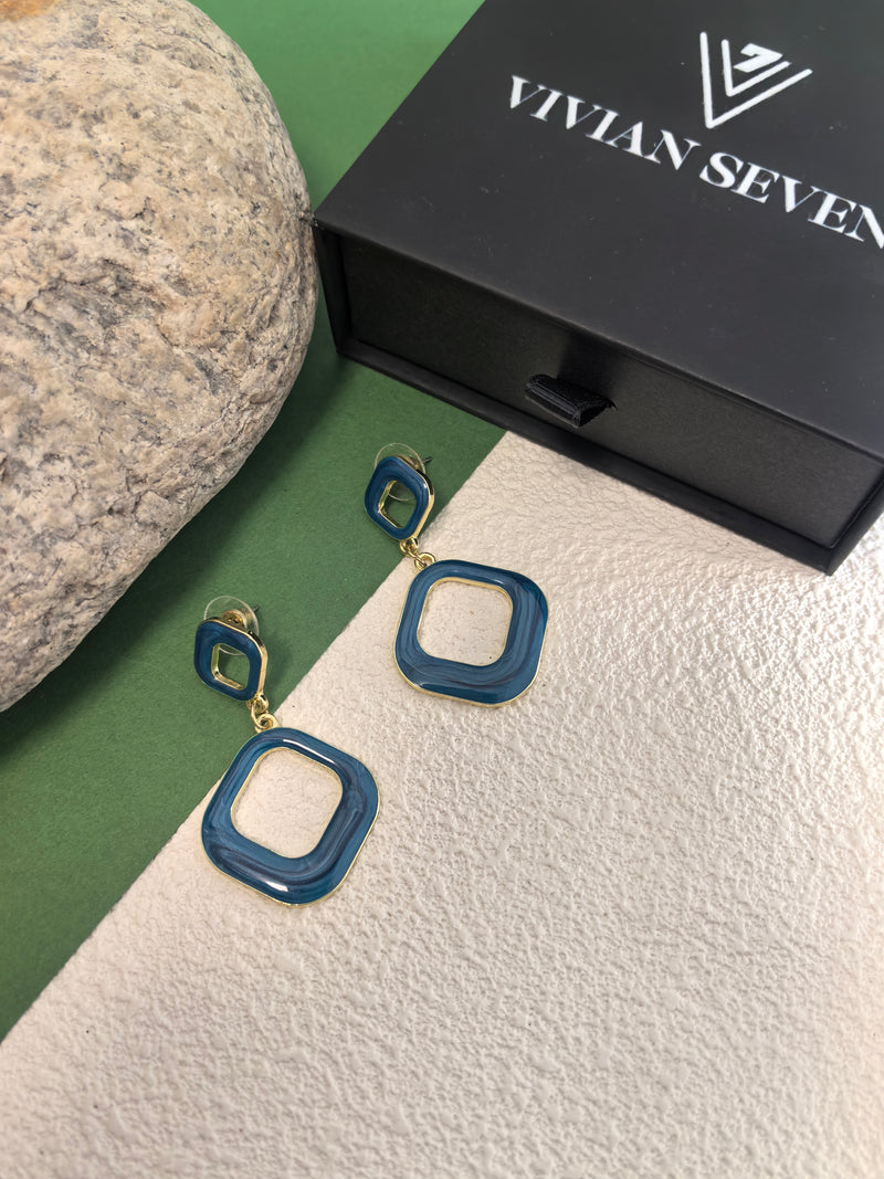 A pair of blue enamel earrings displayed on a beige and green background. The earrings are made of two connected squares and are next to a black box with the brand name "VIVIAN SEVEN."