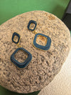 The blue enamel earrings are shown resting on a light-colored rock. The earrings are made of two connected squares and have a gold-tone base.