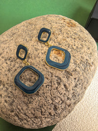 The blue enamel earrings are shown resting on a light-colored rock. The earrings are made of two connected squares and have a gold-tone base.