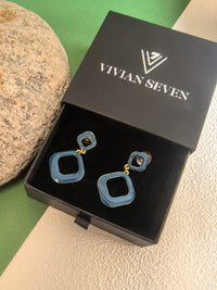  A pair of blue enamel earrings displayed in their black box with the "VIVIAN SEVEN" logo. The background is a mix of beige and green.
