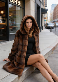 Model sitting on street curb wearing Vivian Seven brown faux fur winter coat with hood over head.