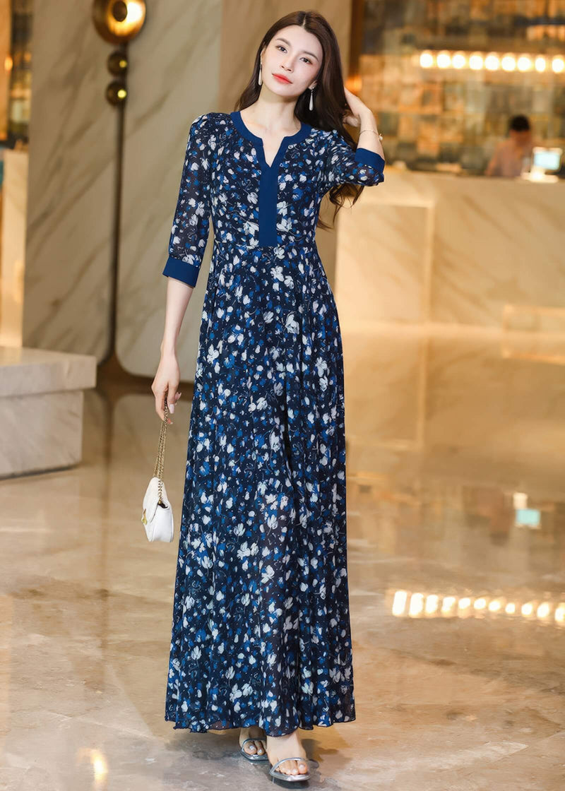  A three-quarter length pose of a woman in a navy floral maxi dress, holding a white quilted clutch bag against her hip. She is wearing elegant drop earrings and silver heels.