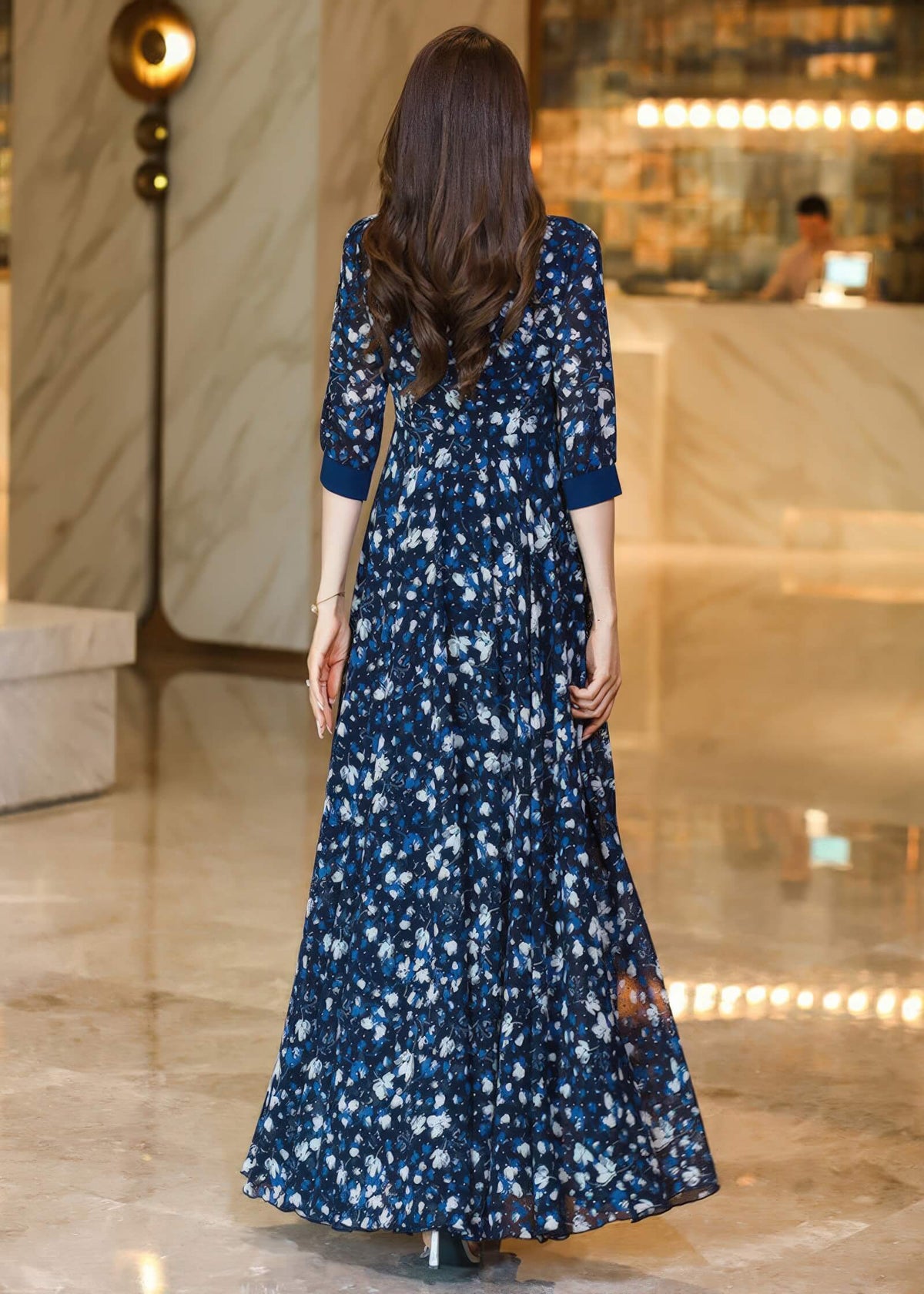  The back view of a woman wearing a navy blue floral maxi dress. The dress features a fitted waist and a flowing, floor-length skirt. Her long, wavy brown hair falls down her back.
