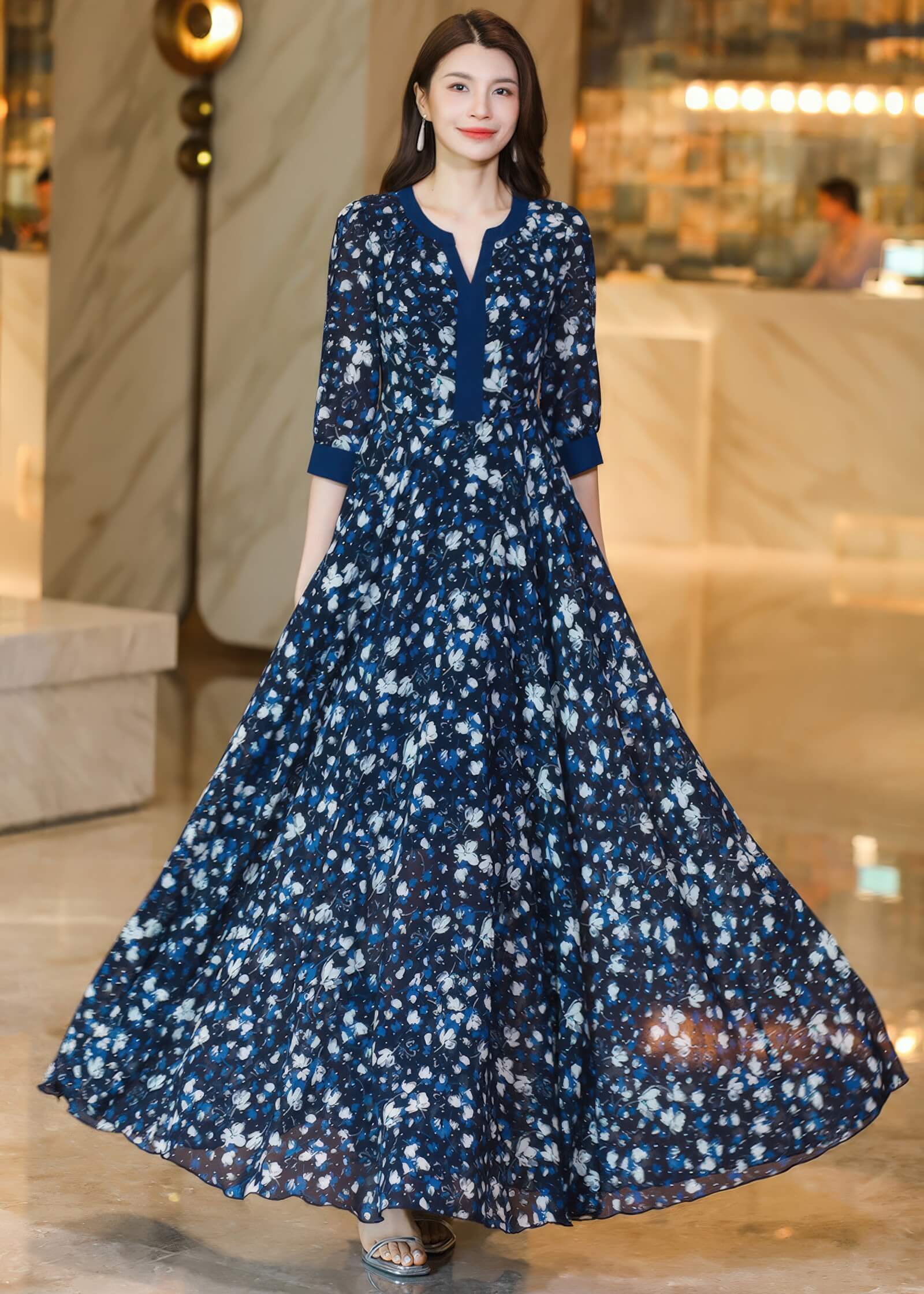 A woman stands facing forward in a navy blue floral maxi dress, running her hand through her hair. The dress has a distinctive navy blue rectangular panel at the V-neckline.