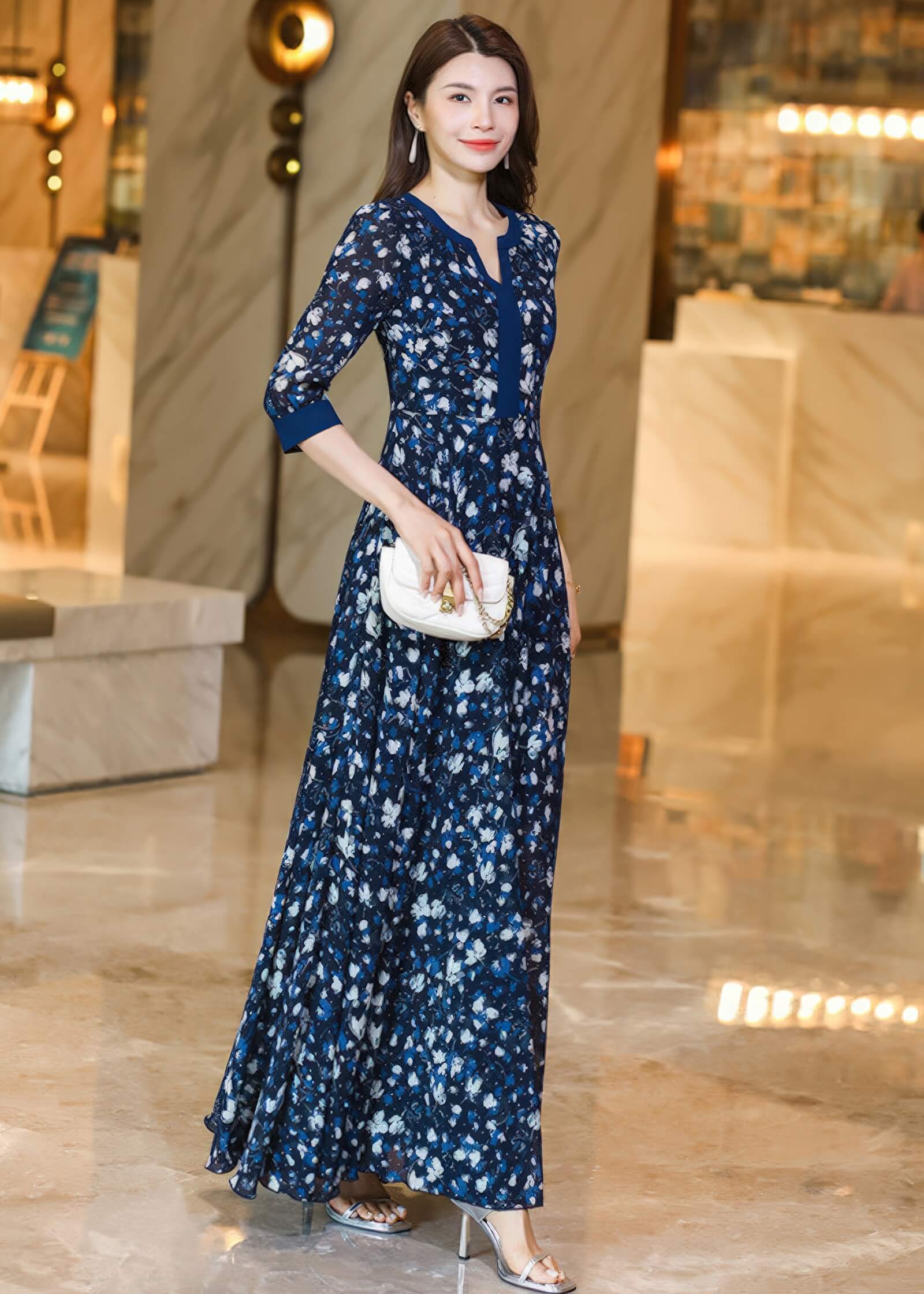 A woman posing in a navy floral maxi dress, holding a white purse in one hand and touching her hair with the other. The background shows a blurry, upscale indoor setting with warm lighting.