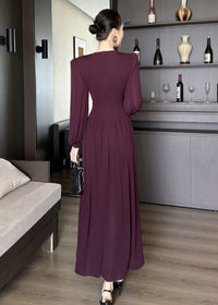 Full-length rear view of a woman standing and wearing a long-sleeved, floor-length maxi dress in a deep burgundy color. The dress features a fitted waist, gathered styling at the lower back, and a discreet zipper closure. She is wearing her hair in a sleek bun and holding a black quilted handbag.