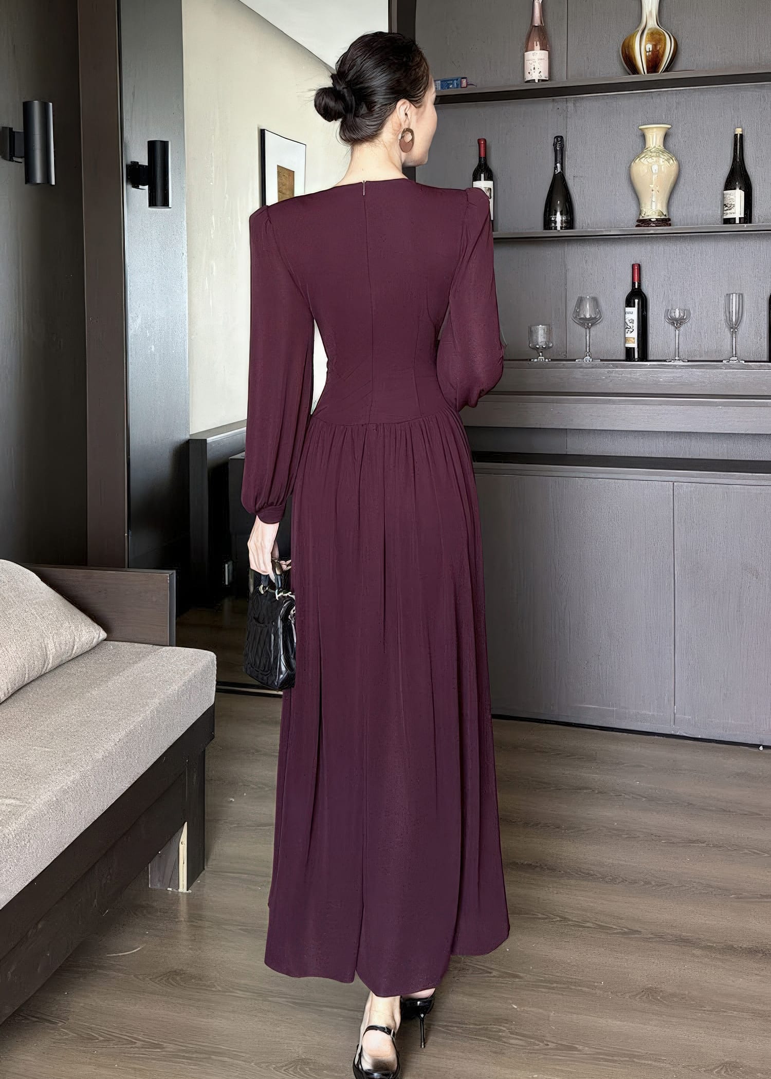 Full-length rear view of a woman standing and wearing a long-sleeved, floor-length maxi dress in a deep burgundy color. The dress features a fitted waist, gathered styling at the lower back, and a discreet zipper closure. She is wearing her hair in a sleek bun and holding a black quilted handbag.