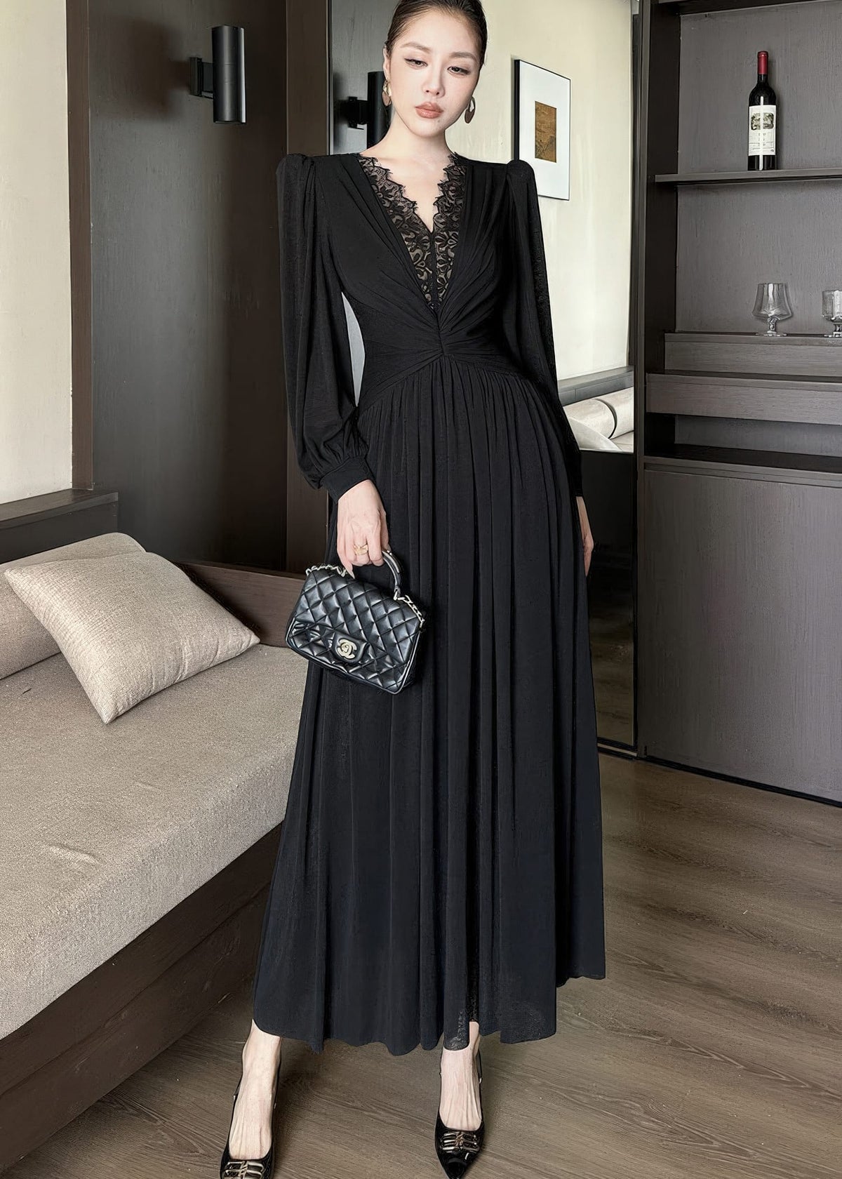 Full-length front view of a woman standing next to a sofa, posing in a black maxi dress. The gown includes long sleeves, a plunging V-neck with black lace patchwork, a ruched bodice, and a pleated maxi skirt. She is holding a black quilted handbag.