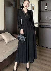 Full-length front view of a woman standing next to a sofa, posing in a black maxi dress. The gown includes long sleeves, a plunging V-neck with black lace patchwork, a ruched bodice, and a pleated maxi skirt. She is holding a black quilted handbag.