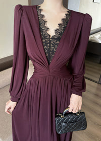 Close-up shot focusing on the bodice of a burgundy maxi dress. The image highlights the plunging V-neckline inlaid with detailed black lace patchwork, the fine ruching and twisted knot styling at the waist, and the gathered texture of the long sleeves. The woman is holding a black quilted handbag.