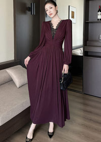 Full-length front view of a woman posing in a burgundy maxi dress. The dress features long bishop sleeves, a plunging V-neckline inlaid with black lace patchwork, a twisted knot detail at the waist, and a flowing skirt. She is holding a black quilted handbag and wearing black heels.